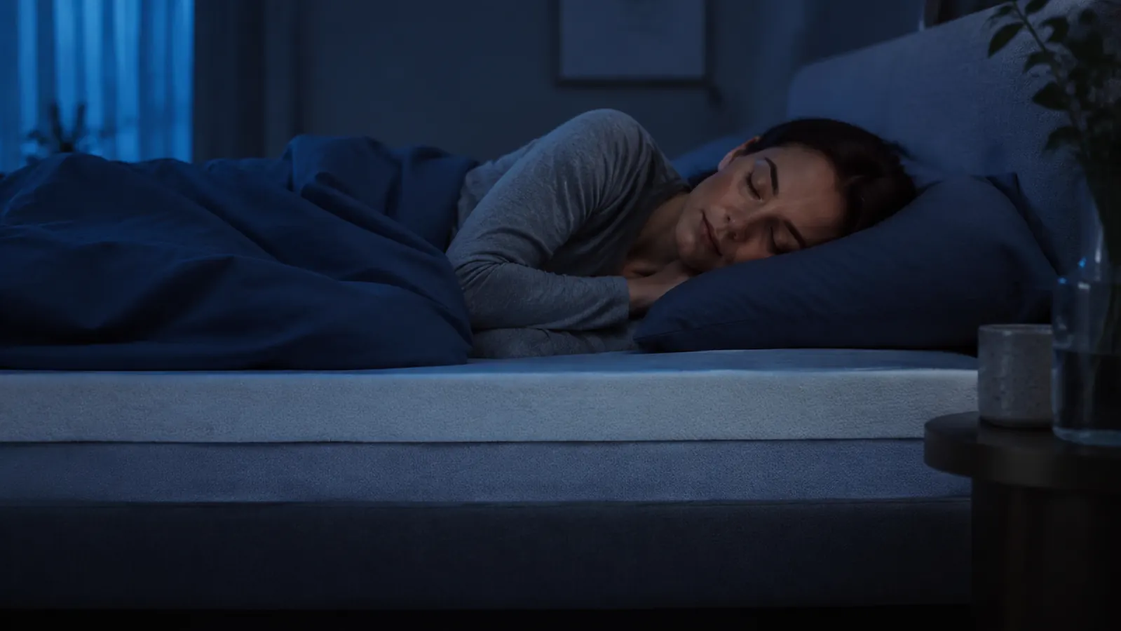 Person sleeping comfortably on their side in a cool dark bedroom, relaxed posture and layered bedding visible, suggesting deep restful sleep