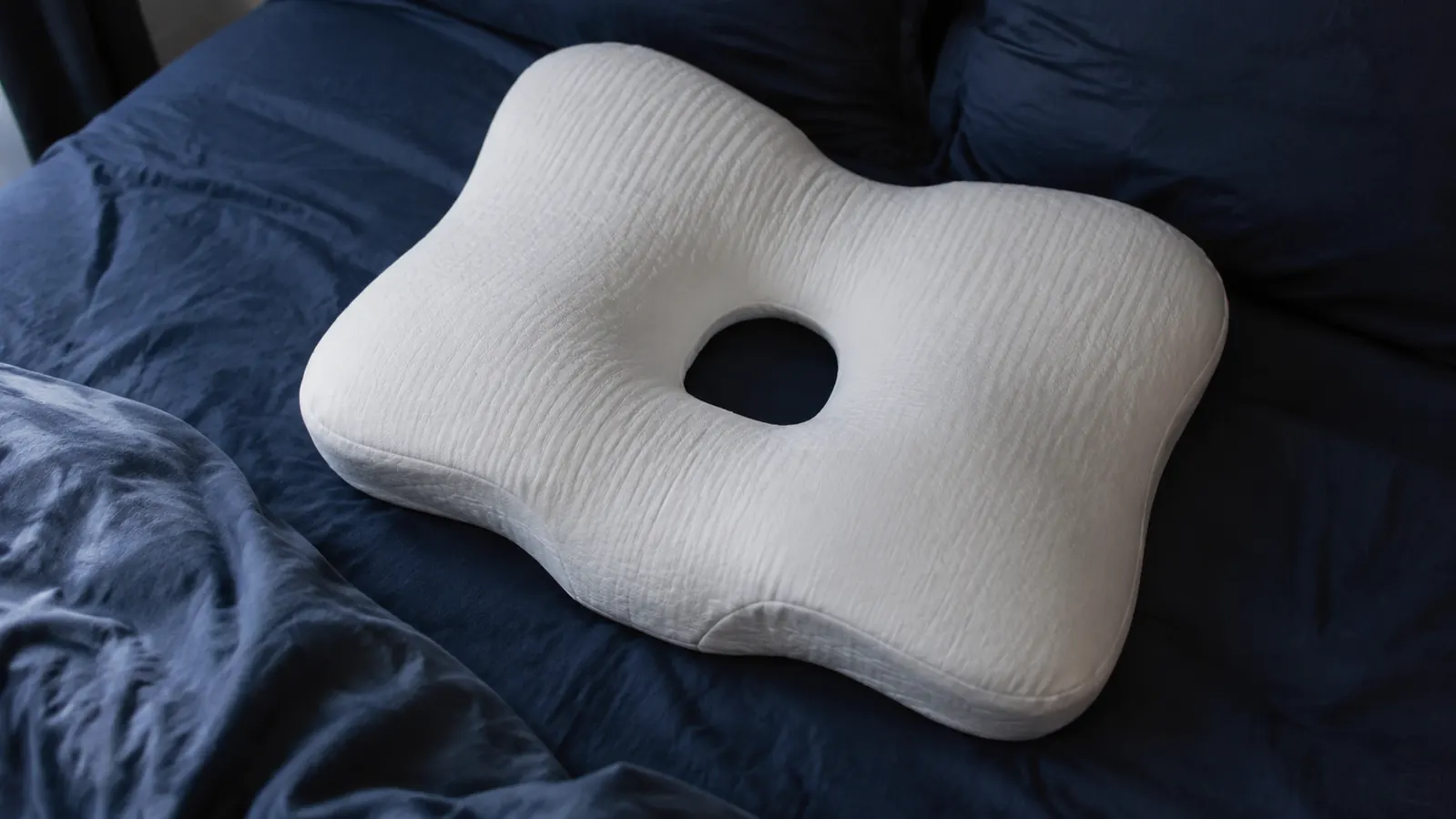 Close-up of the Osteo Cervical Pillow on a bed showing the contoured dual-lobe shape and hollow center against dark navy bedding